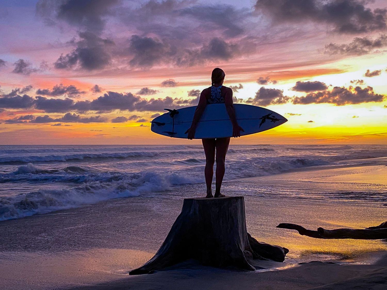 Silhouetted surfer stands on beach stump holding surfboard against vibrant purple and orange sunset sky over ocean waves.