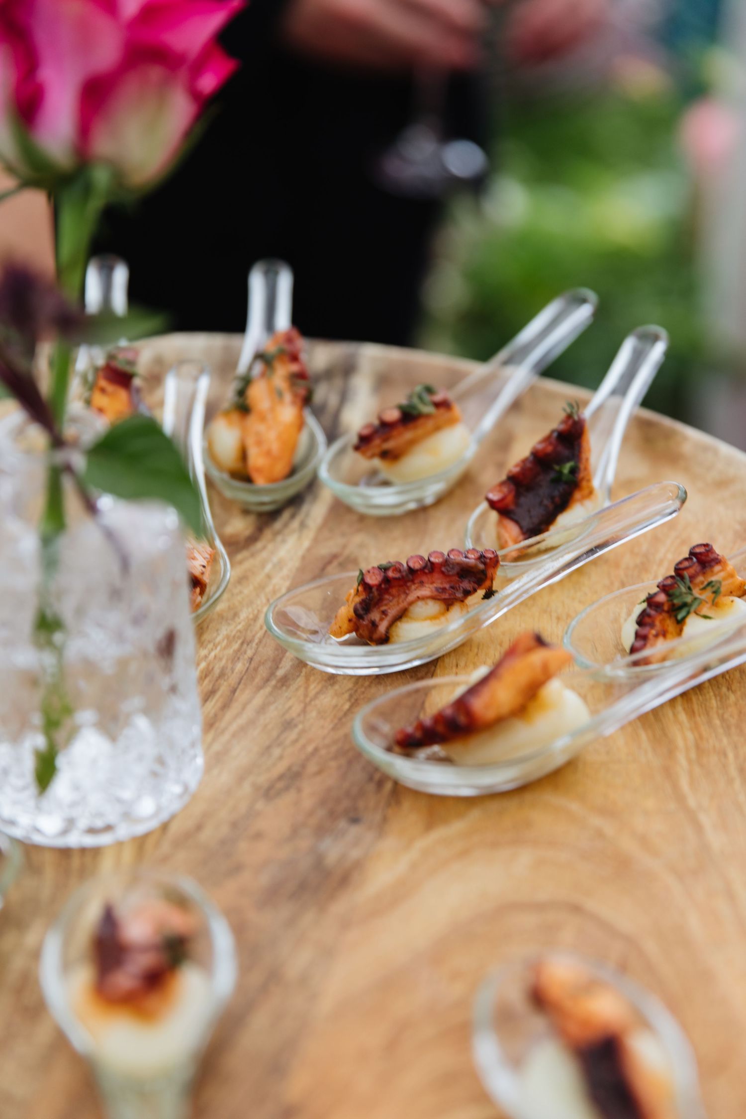 Elegant appetizers served on small spoons displayed on wooden board with pink flowers.