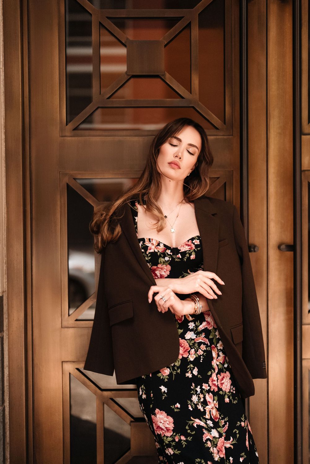 Stylish woman in floral dress and brown blazer poses against ornate wooden door.