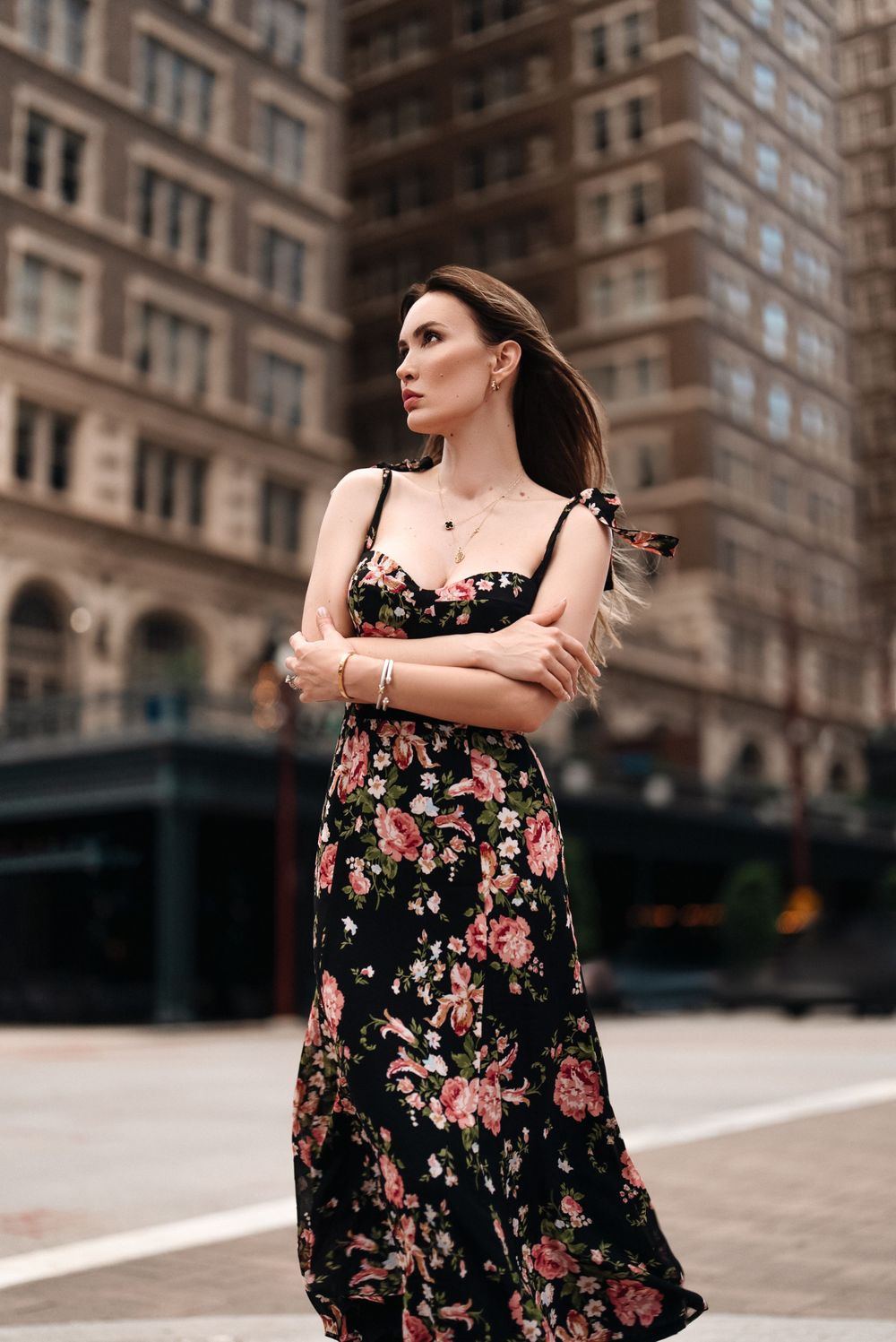 Woman in floral dress stands confidently in front of urban building with arms crossed.