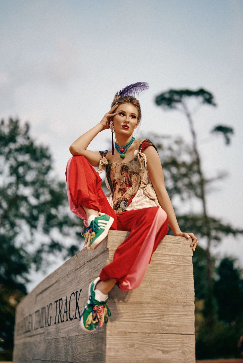A model in red pants and colorful attire poses artistically on a wooden surface outdoors.
