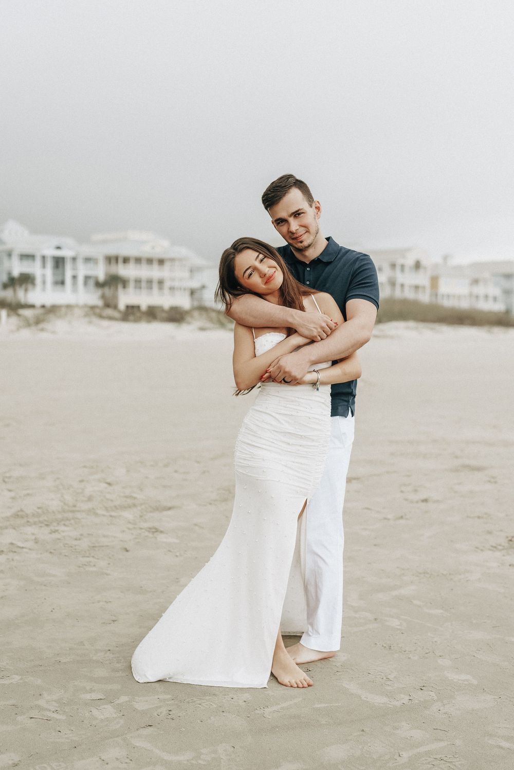 Romantic couple embraces on sandy beach with coastal homes in background.