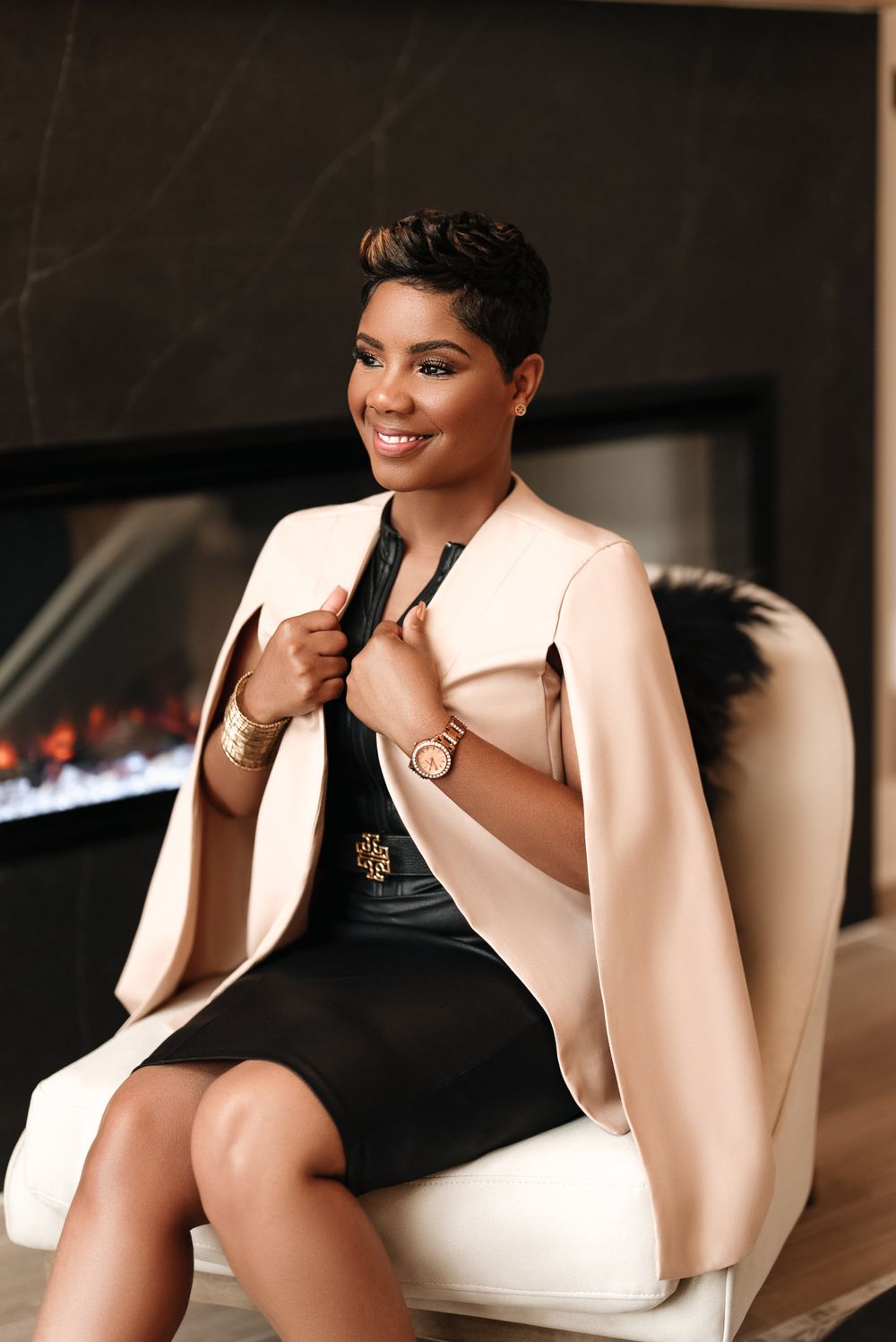 Professional woman in cream blazer and black dress sits elegantly by fireplace.