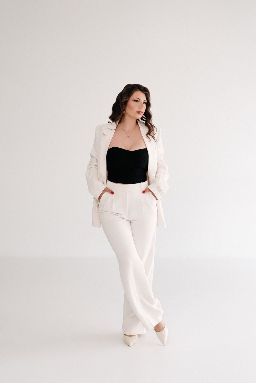 Fashion model poses in white pantsuit with black top against minimalist background.