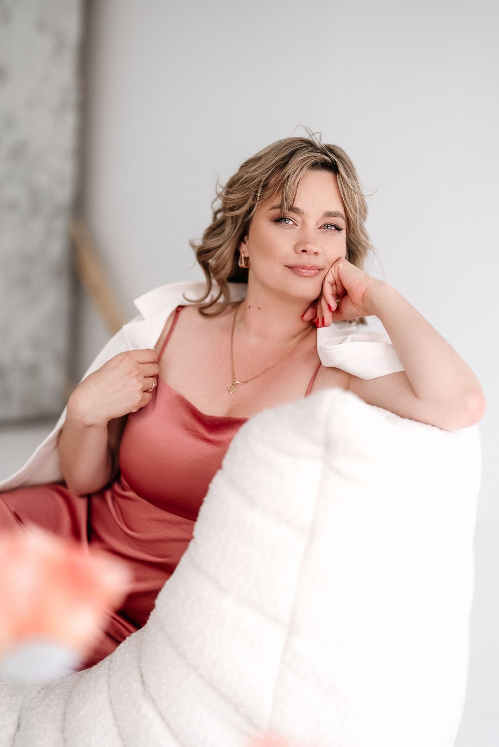 Woman in coral slip dress reclines on white couch in bright studio setting.