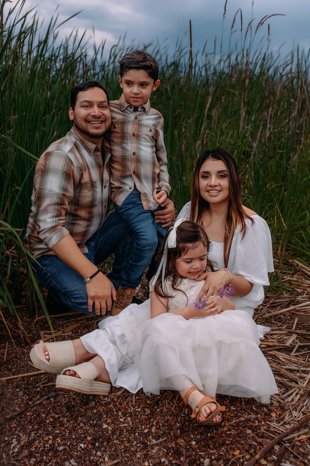 Family in coordinating plaid and white outfits sit together on a rocky beach surrounded by tall grass at sunset.