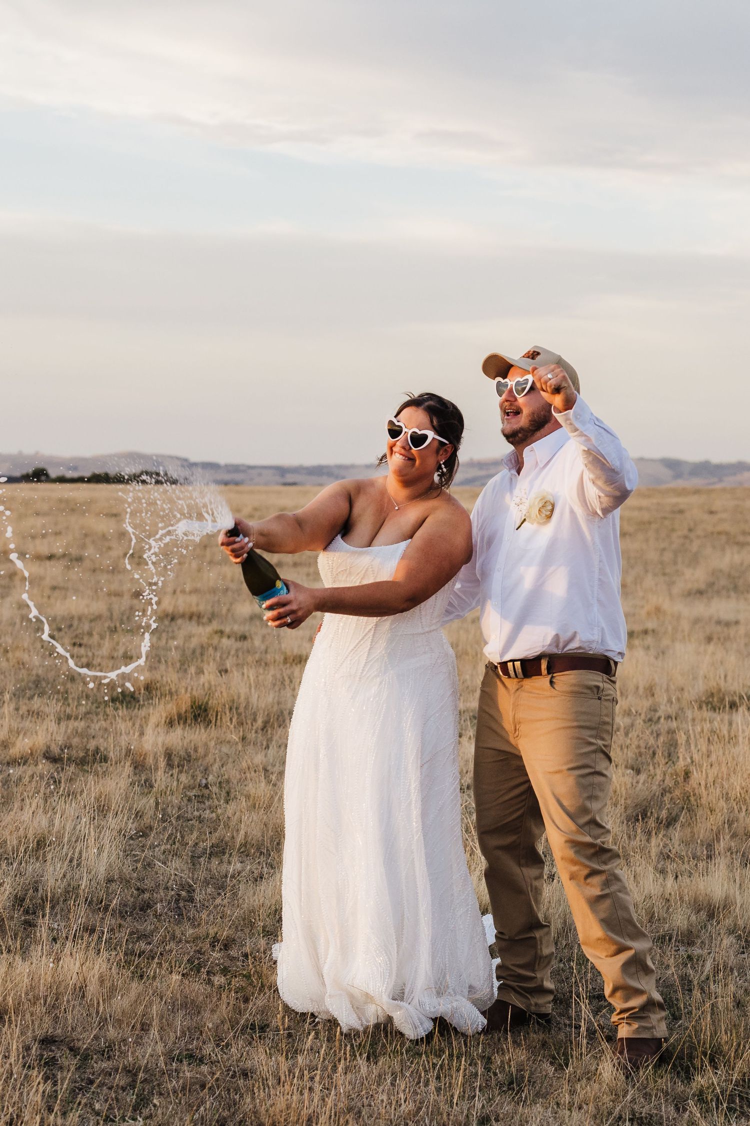 A couple in wedding attire celebrates by popping champagne in a golden field at sunset.