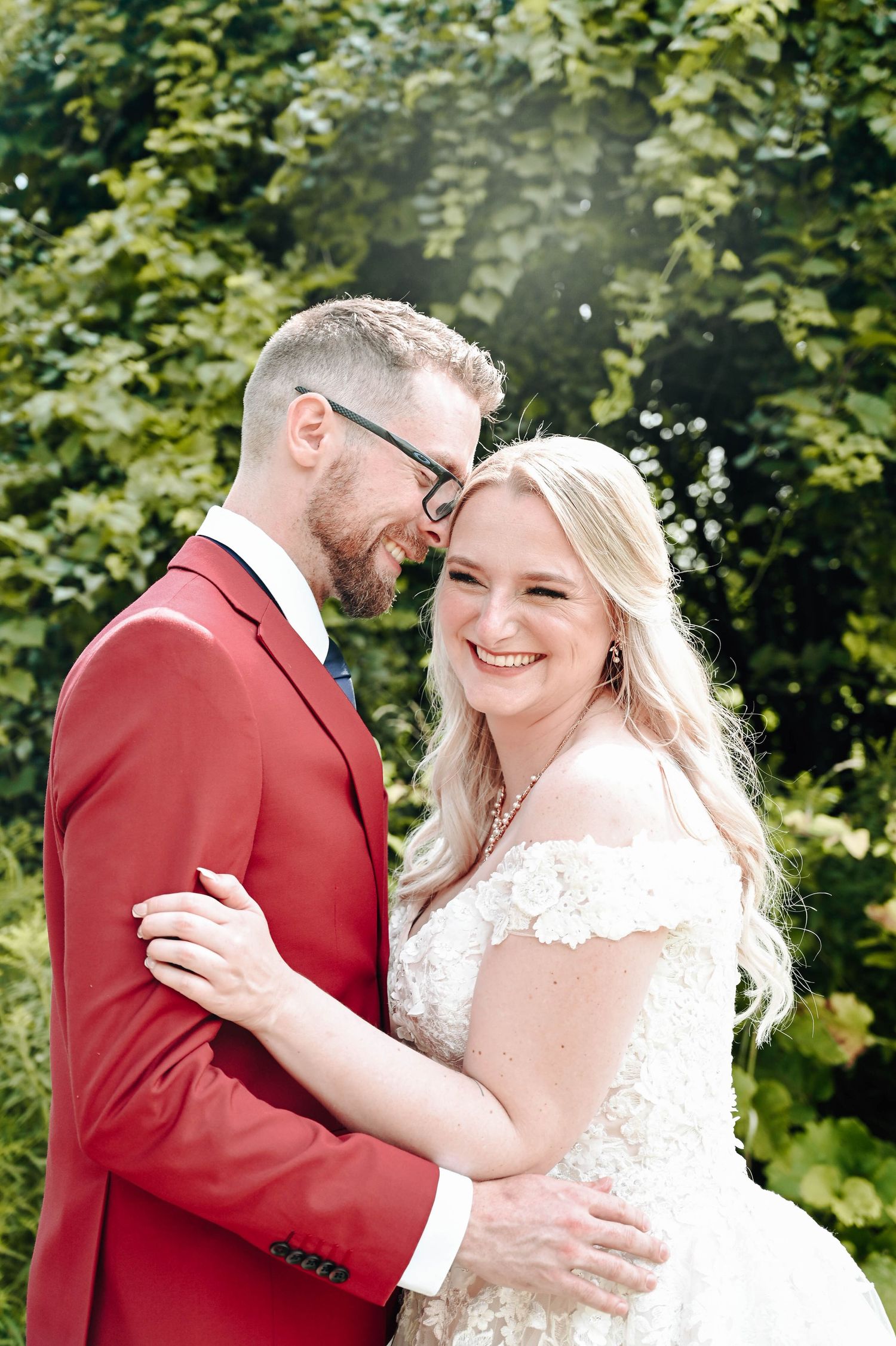 A joyful wedding couple embraces outdoors, with the bride in white lace and groom in a burgundy suit against green foliage.