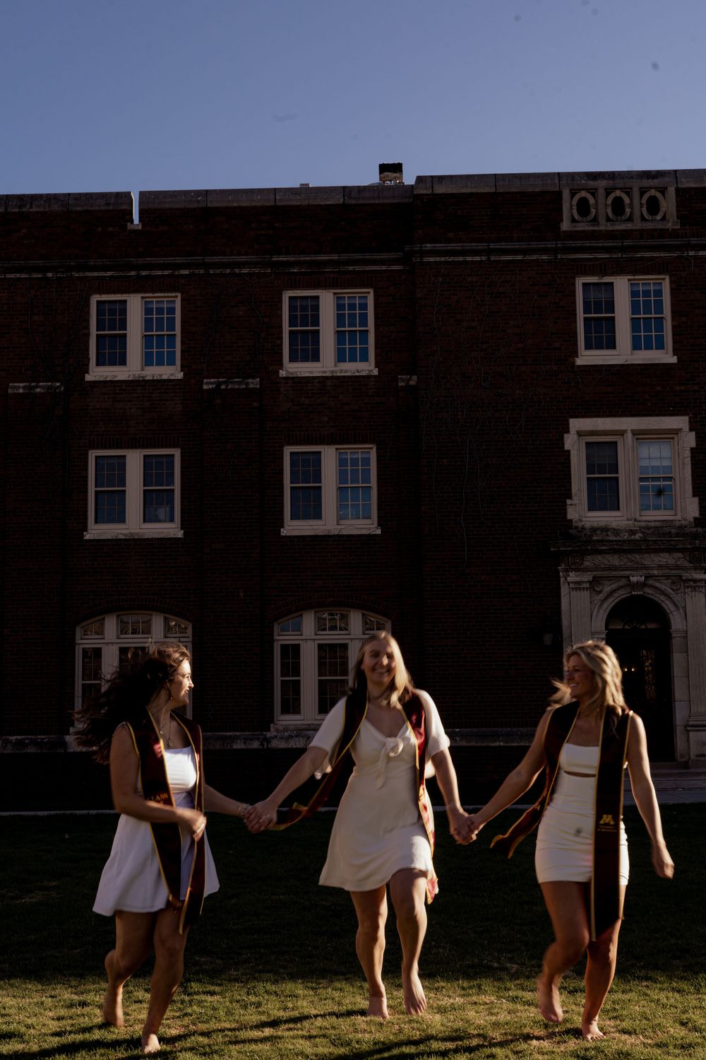 Three graduates in white dresses walking hand in hand on campus lawn at sunset.