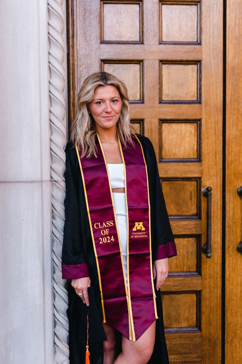 Graduate wearing maroon stole and black gown poses in front of wooden door.