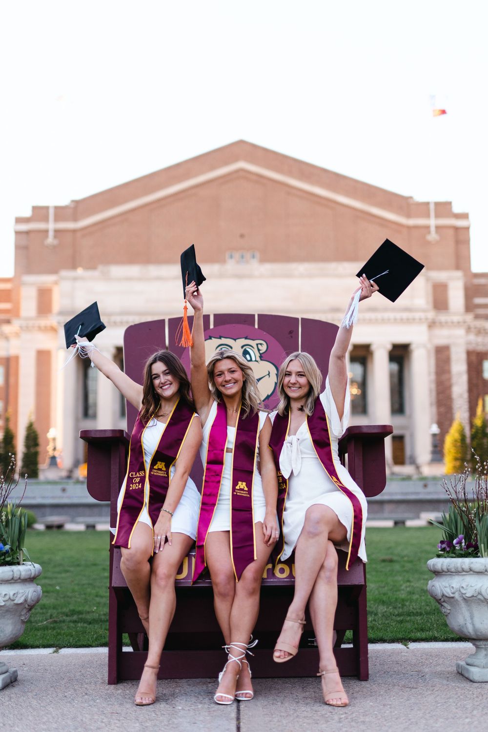 Three graduates in maroon stoles celebrate on large chair outside university building.
