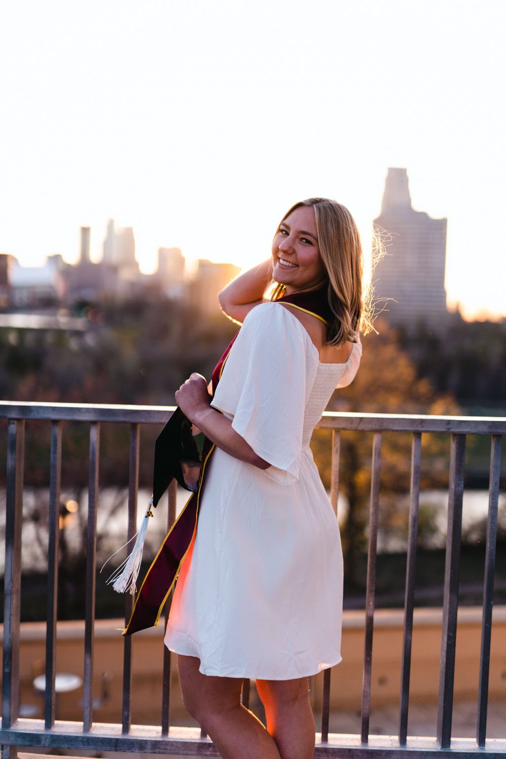 Graduate in white dress poses against city skyline at sunset.