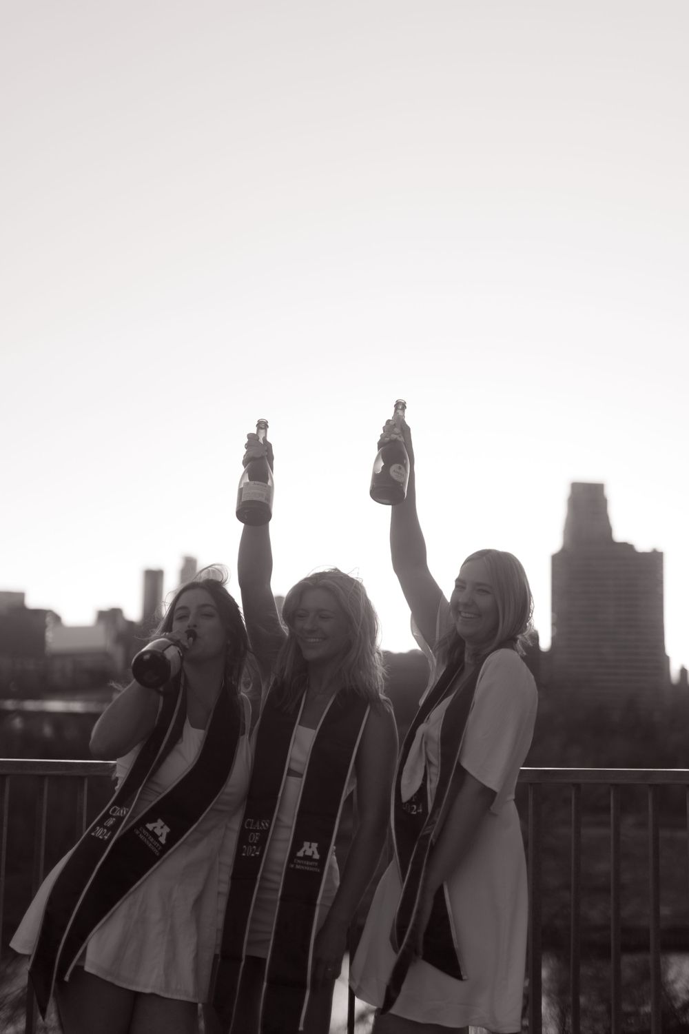 Three graduates celebrate by raising their arms against a city skyline in a black and white photo.