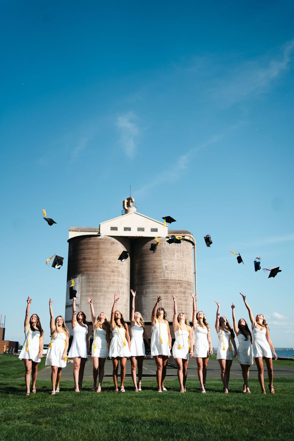 Graduates in white dresses toss caps in celebration in front of industrial grain silos.