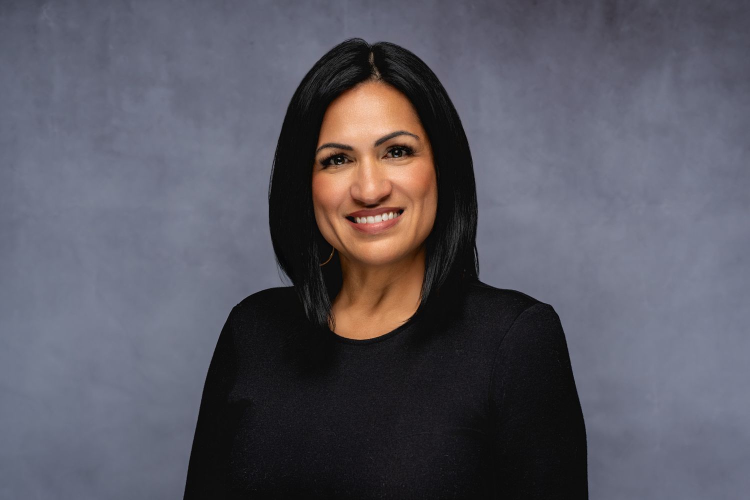 Professional headshot portrait of someone with long dark hair wearing a black top against a gray backdrop while smiling warmly.