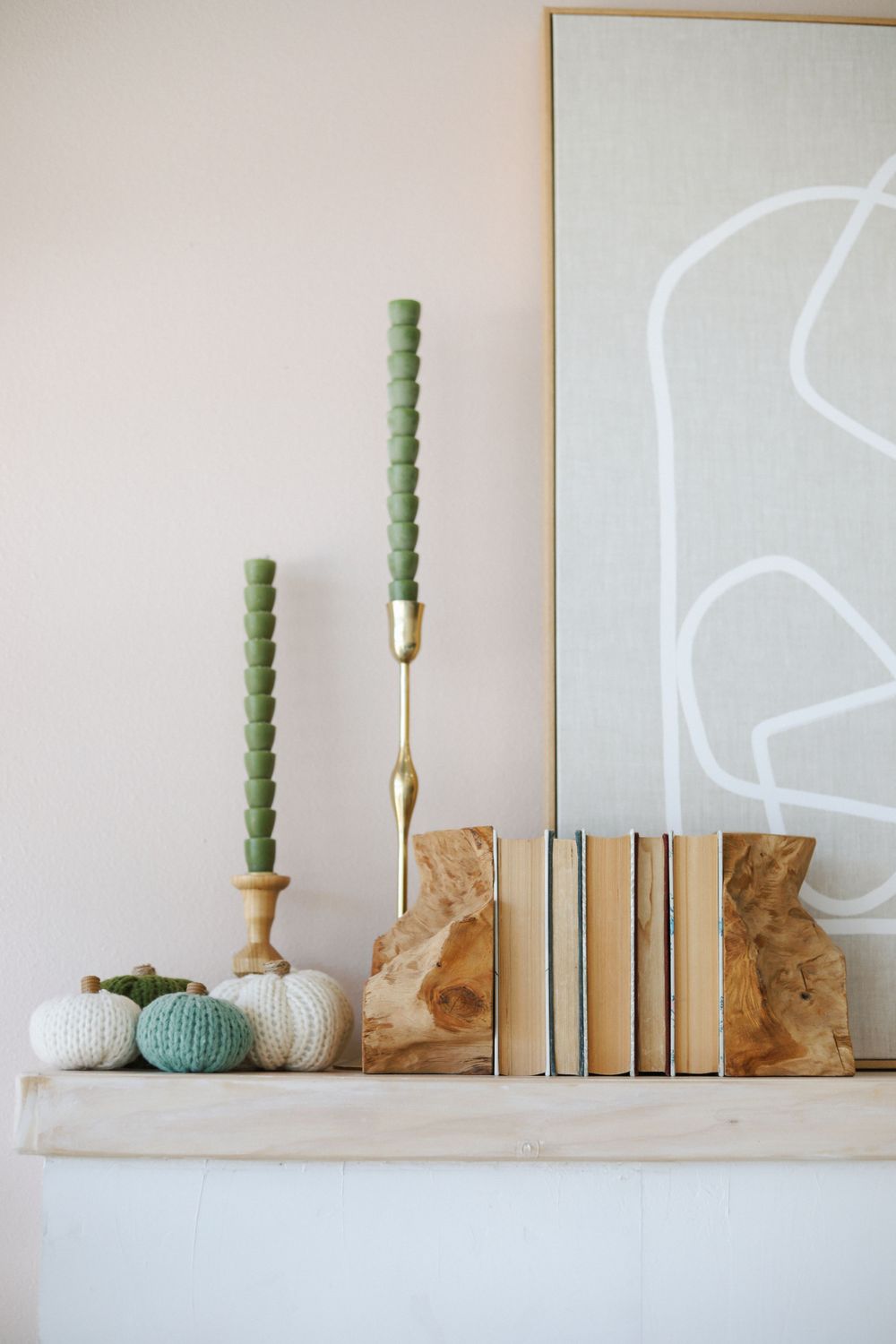 Minimalist shelf display featuring a green candlestick, gold holder, books, and decorative pumpkins against white wall.