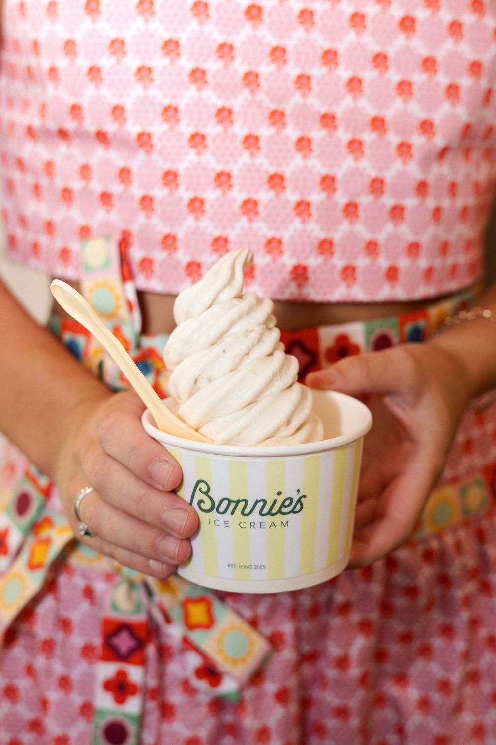 Colorful frozen yogurt in branded cups with wooden spoons against a pink patterned background.