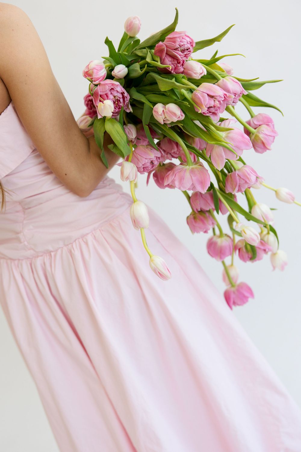 Woman in pink dress holding a large bouquet of pink tulips cascading downward against a white background.