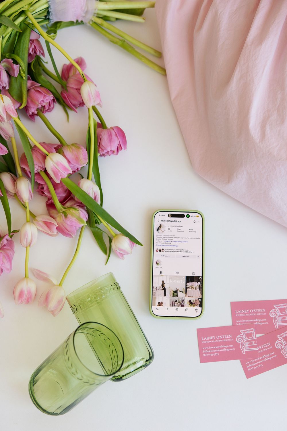 Flat lay of pink tulips, green glass, smartphone showing Instagram, business cards, and pink fabric on white surface.