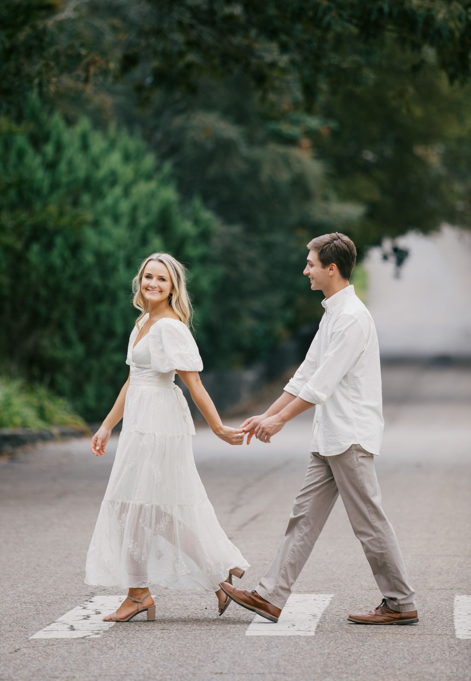A couple in white outfits holds hands while walking across a pedestrian crossing on a tree-lined street.