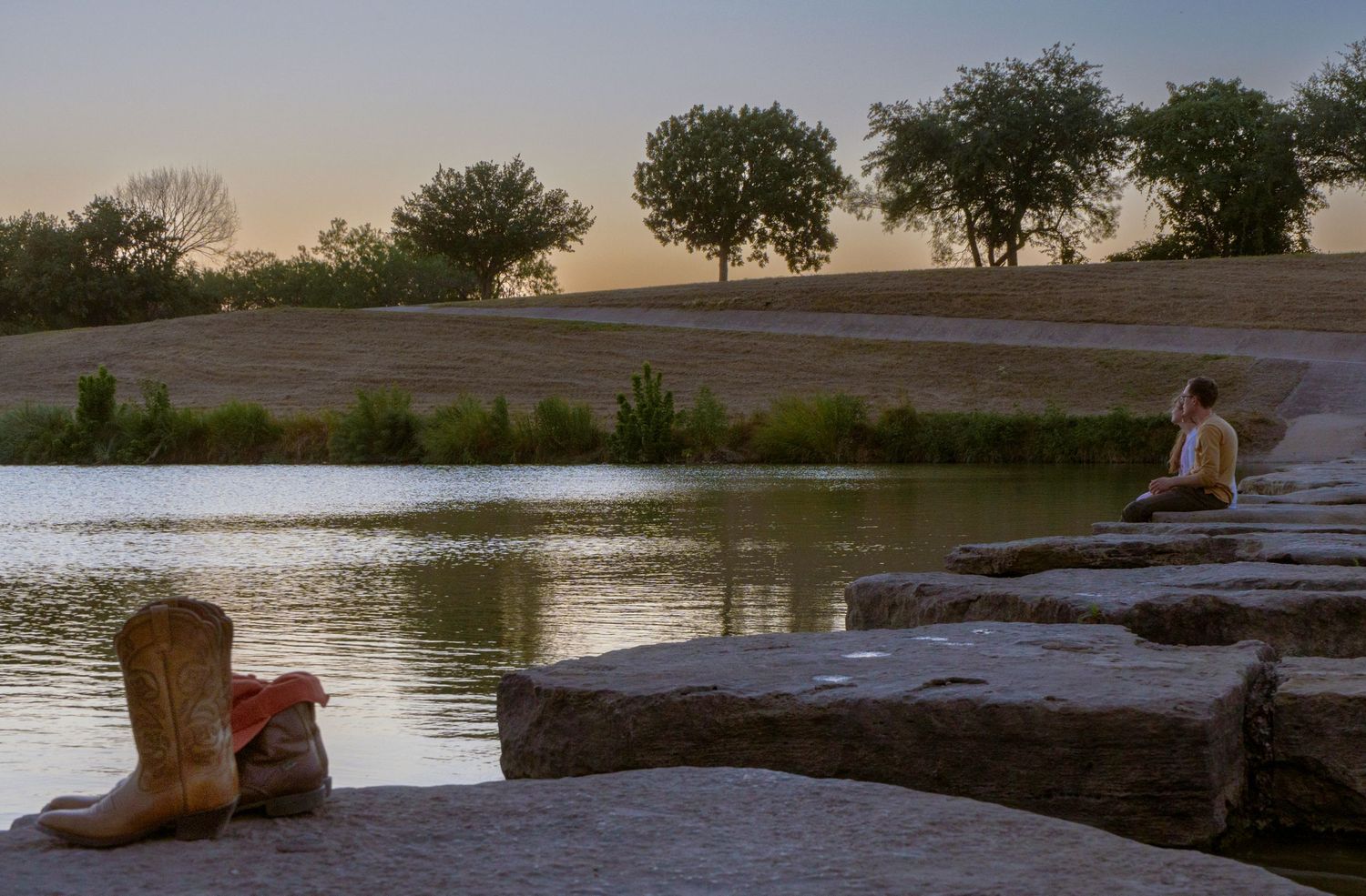 A cowboy boot sits on a rock ledge by a tranquil lake at sunset with trees dotting the hillside in the background.