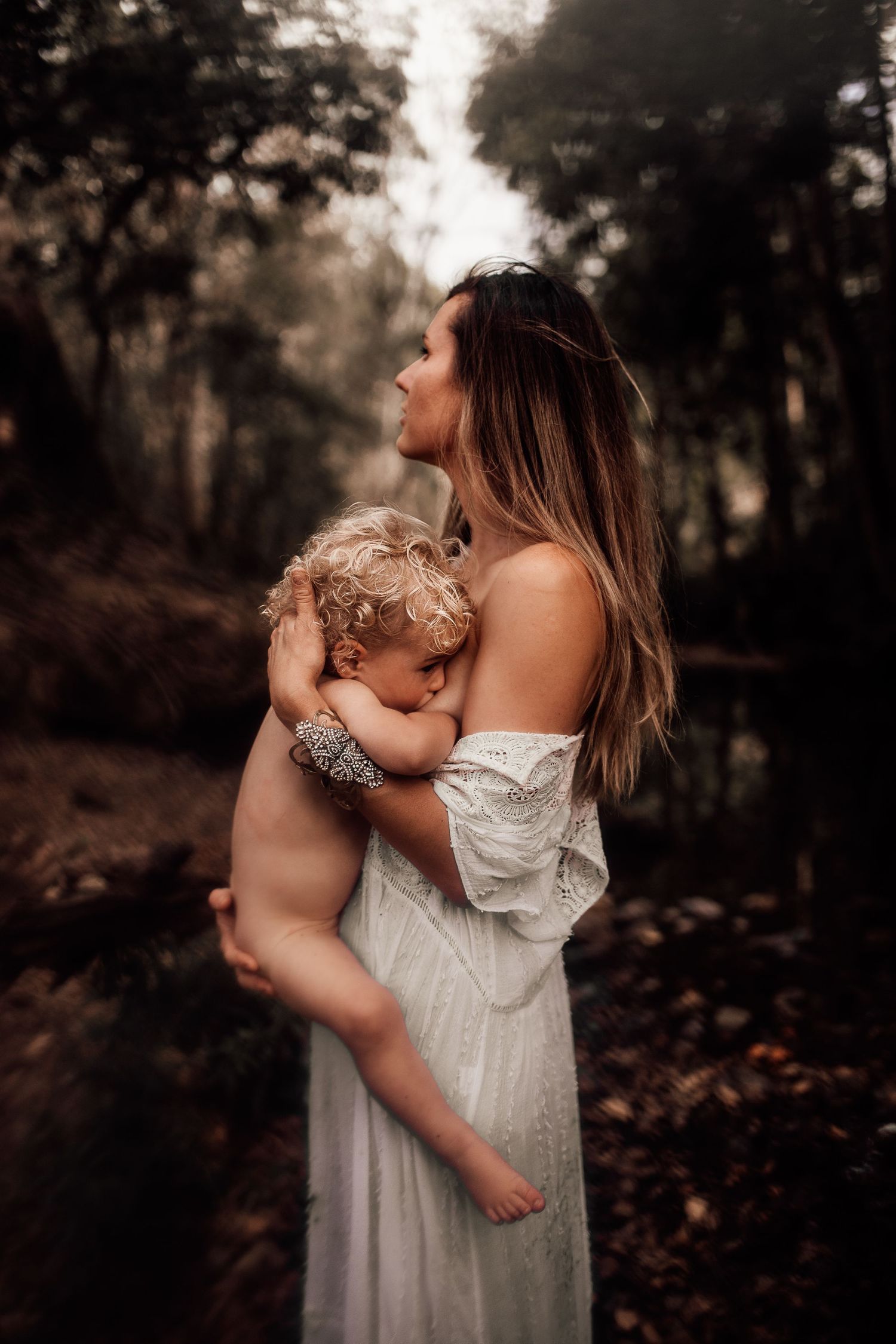 A tender moment between parent and child in a white dress against a moody forest backdrop.