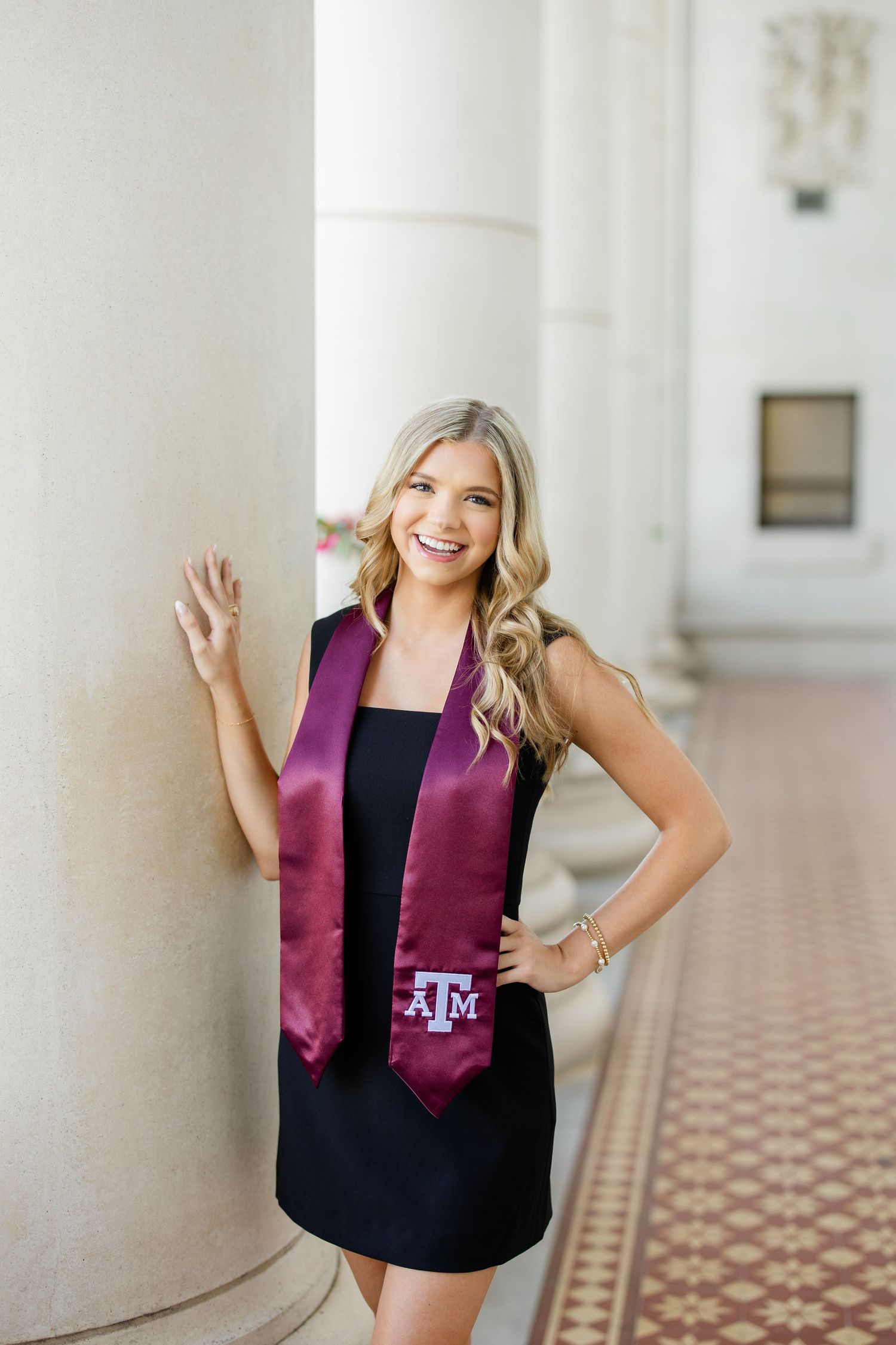 Graduate in black dress and maroon Texas A&M stole poses by white column during graduation photo session.