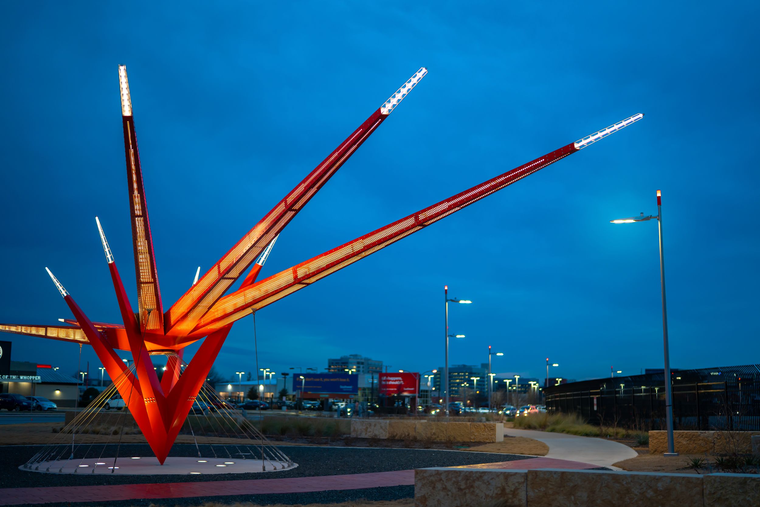 Large red metal sculpture against blue sky at sunset with urban skyline in background.