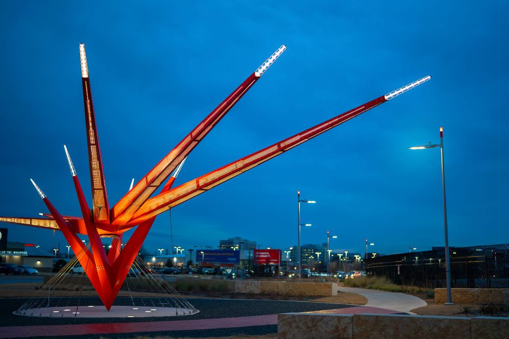 Large red metal sculpture against blue sky at sunset with urban skyline in background.