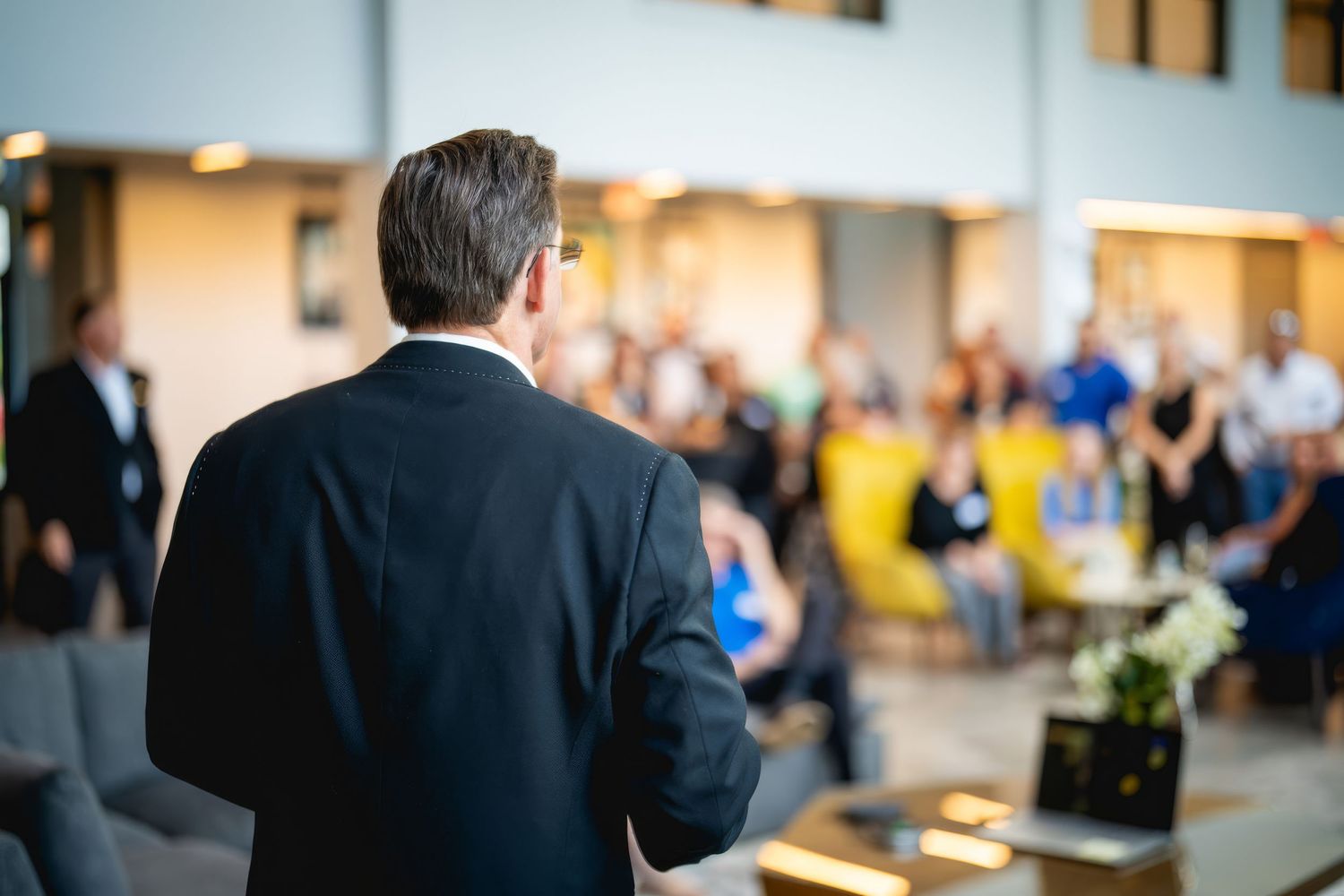 Business speaker in dark suit addressing blurred audience at corporate conference venue.