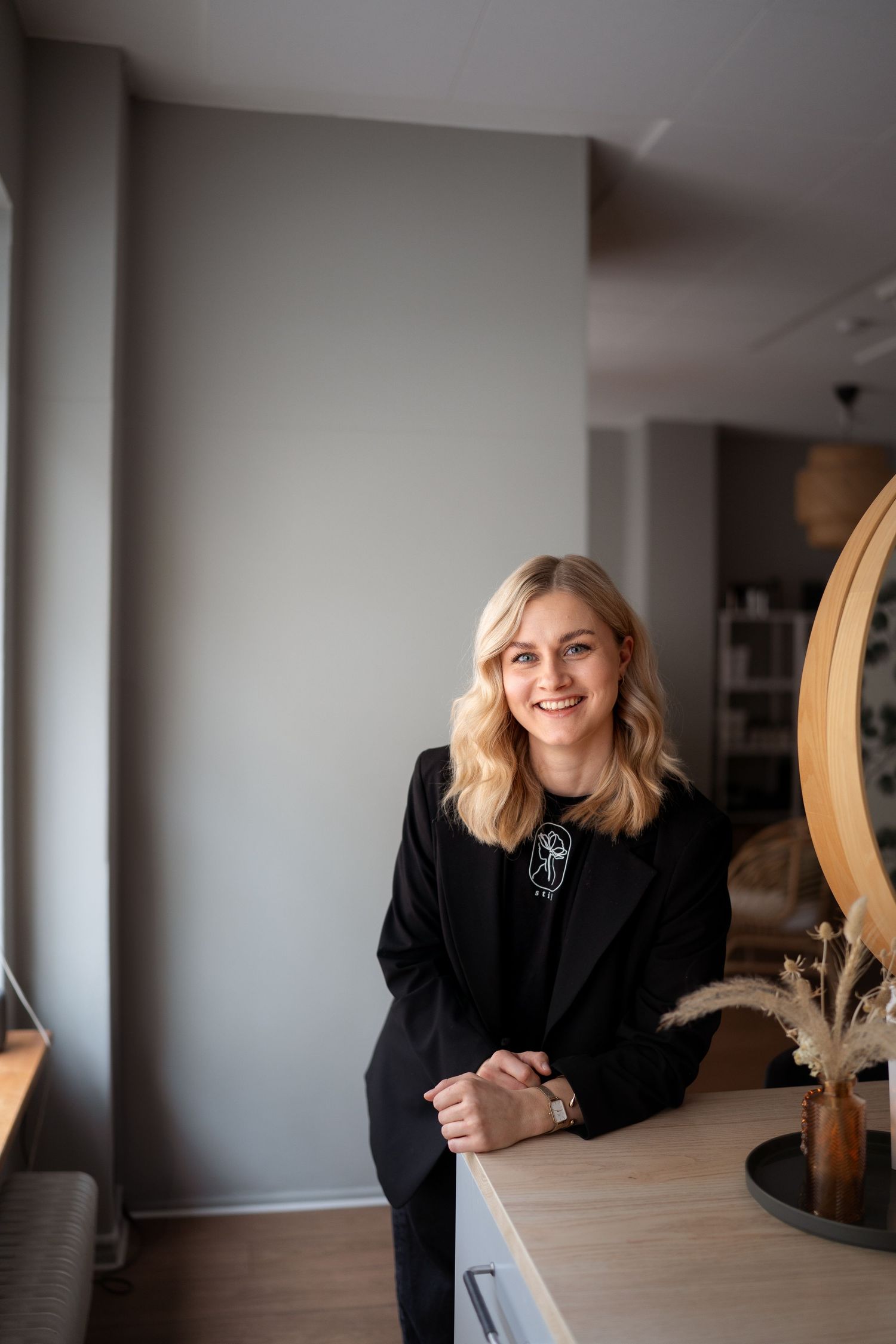Professional in a black blazer sits casually at a wooden table in a modern interior space with natural lighting.
