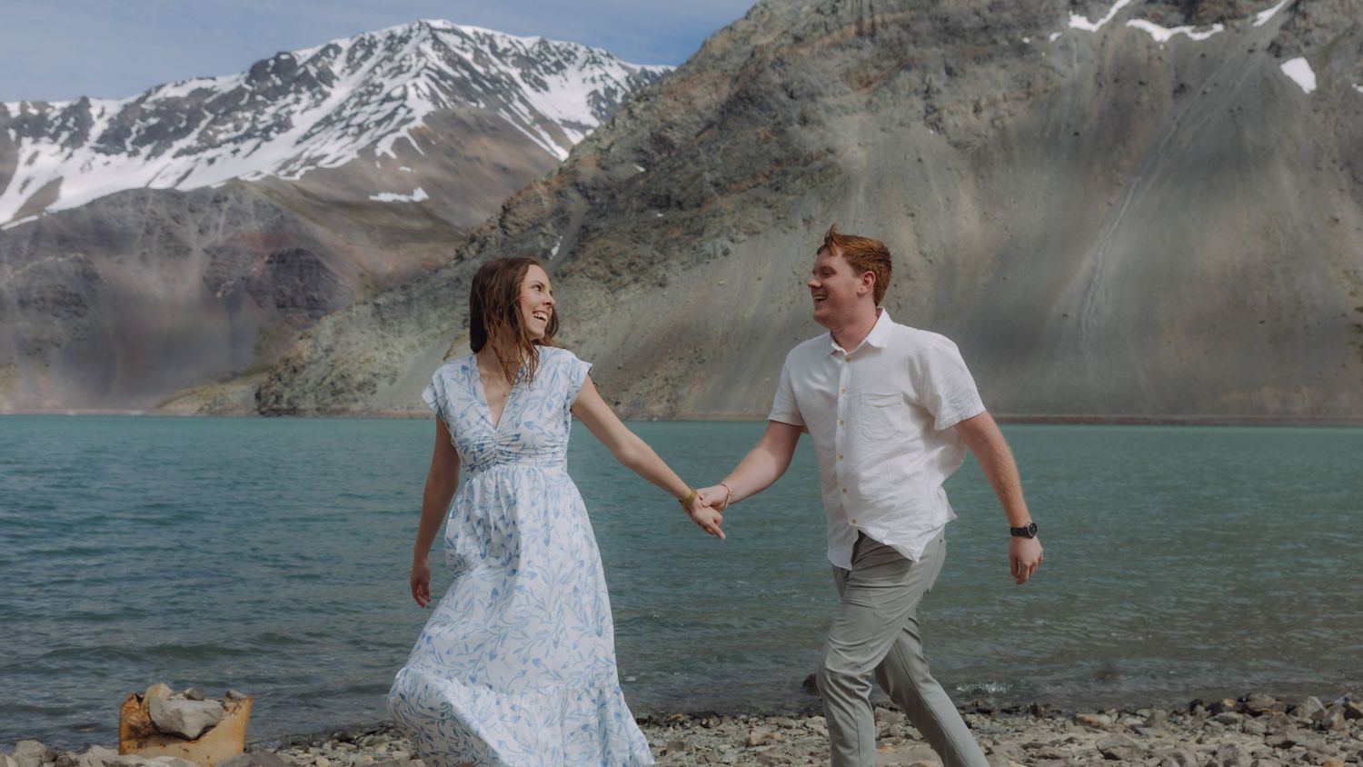 Couple holding hands and walking along rocky shoreline with turquoise glacial lake and snowy mountains behind.