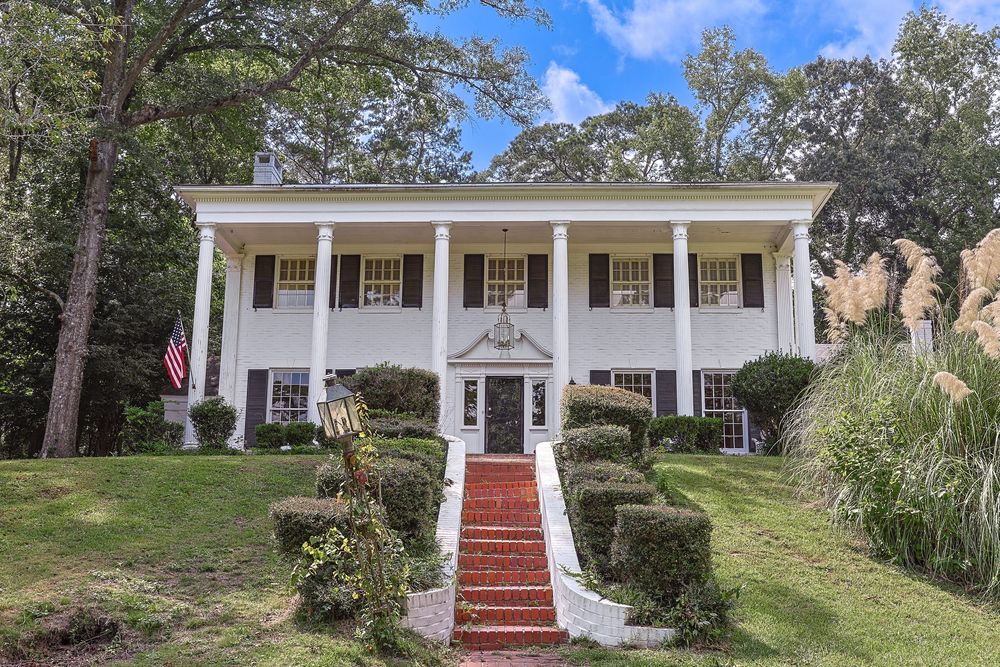 Colonial style home with white columns and red brick steps leading to covered front porch surrounded by landscaped yard.