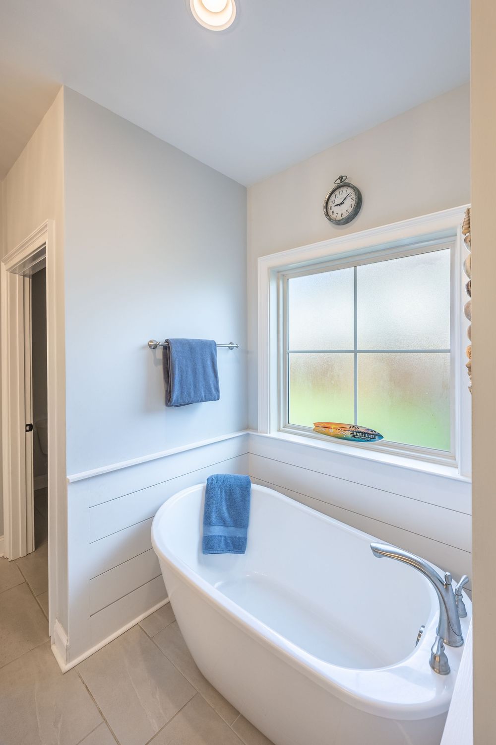 Bright white freestanding soaking tub beside frosted window with blue towels and clock in modern bathroom.