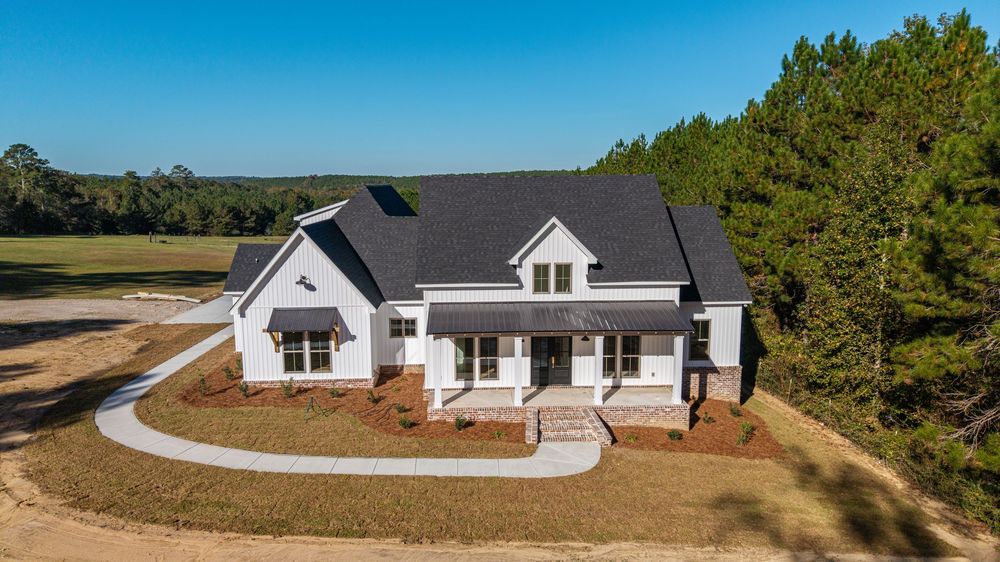 Panoramic drone view of a white modern farmhouse with wraparound porch nestled among pine trees in a rural setting in Ozark, AL.
