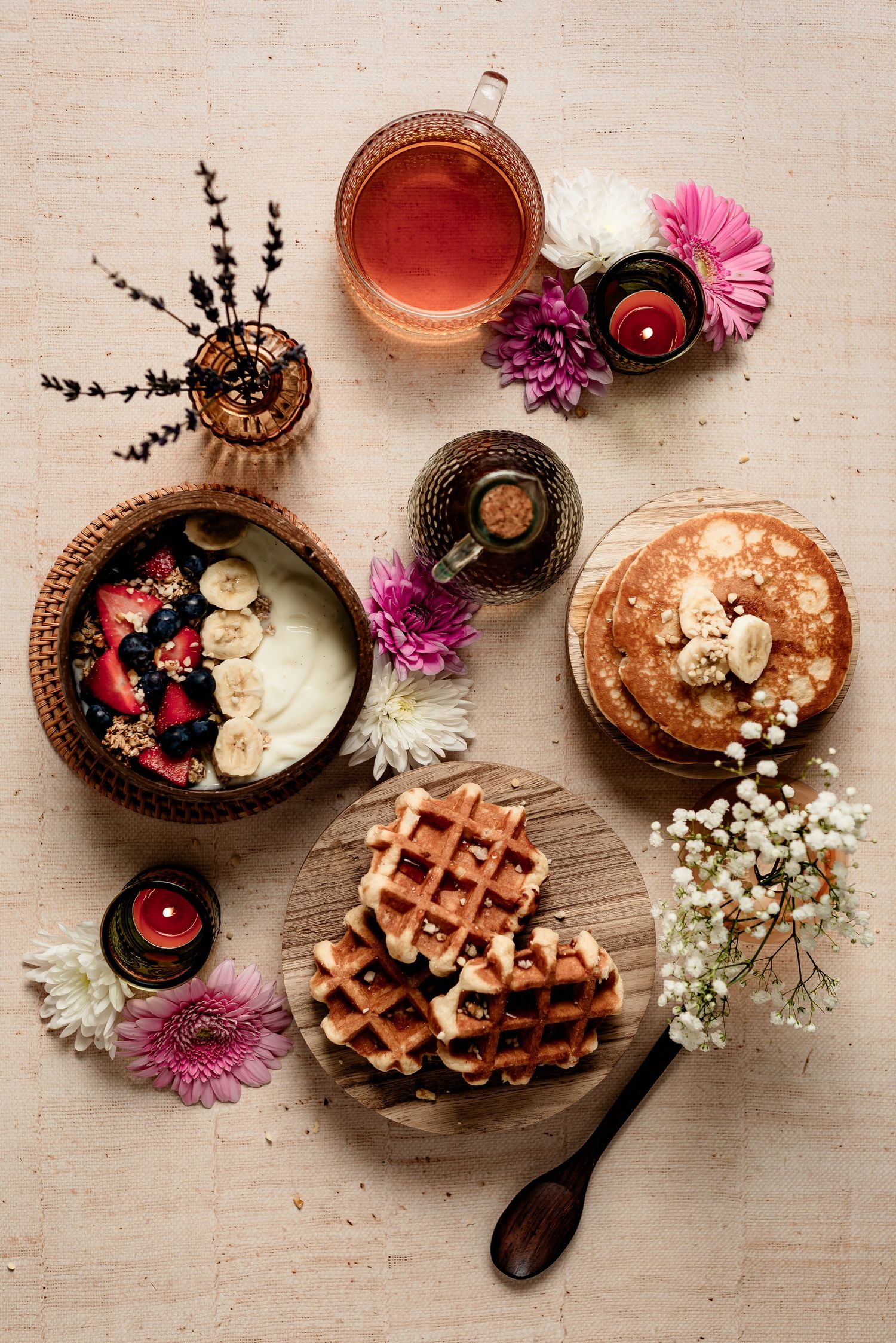 Overhead view of breakfast spread with waffles, flowers, bowls of fruit and yogurt, pink daisies, and herbal tea.