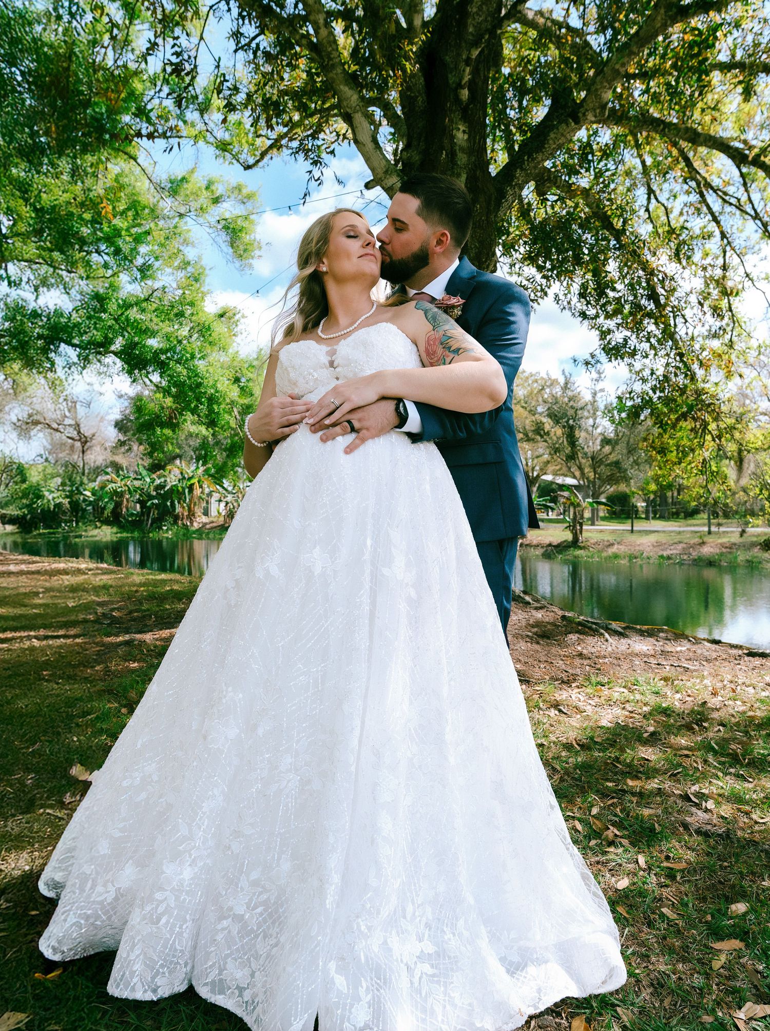 Romantic wedding couple embraces beneath a tree by a tranquil pond on an autumn day.