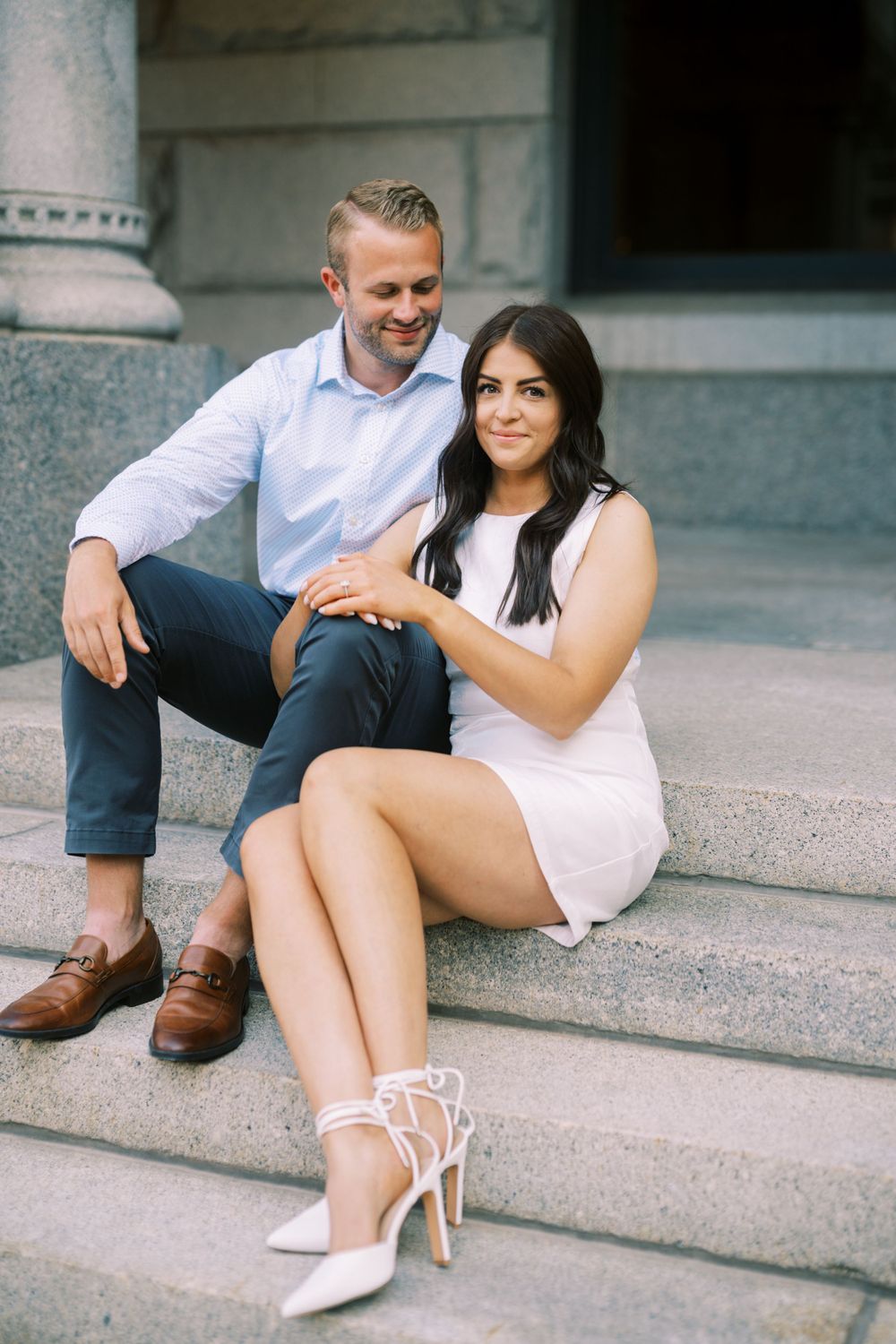 A couple sit together on outdoor stone steps, dressed in light summer attire and white shoes.