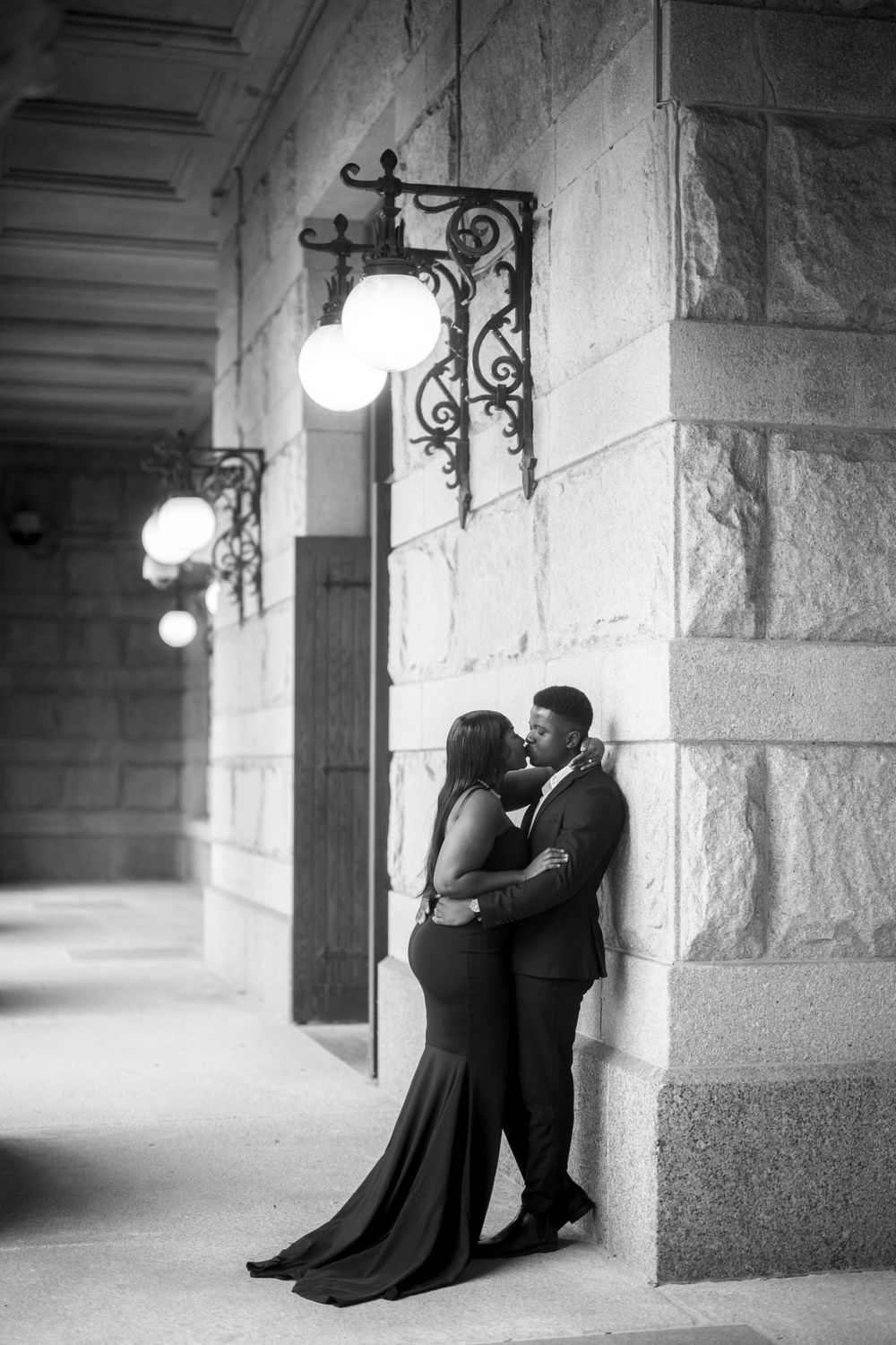 Romantic couple embraces in elegant formal wear under ornate vintage wall sconces in a stone archway corridor.