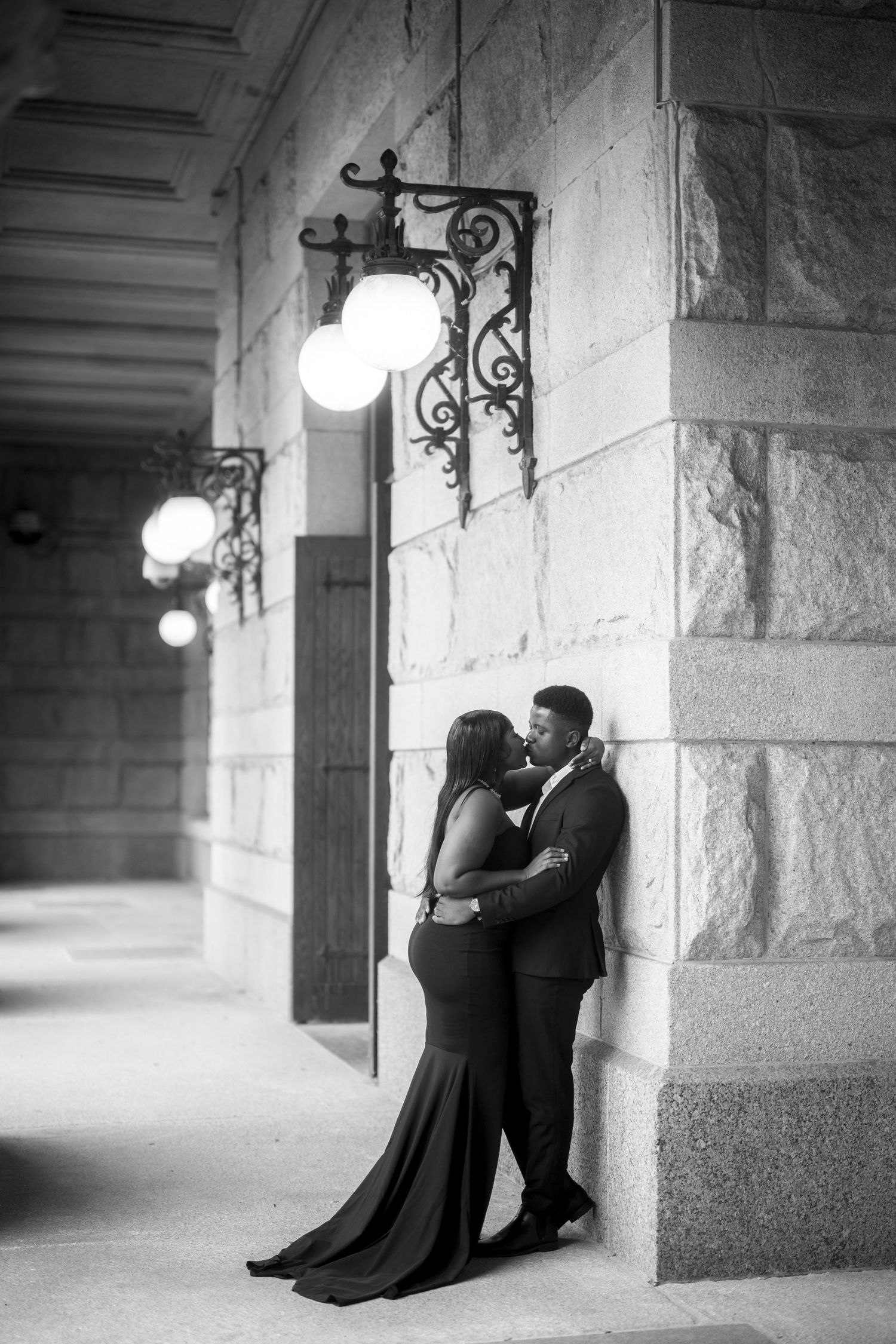 Romantic couple embraces in elegant formal wear under ornate vintage wall sconces in a stone archway corridor.