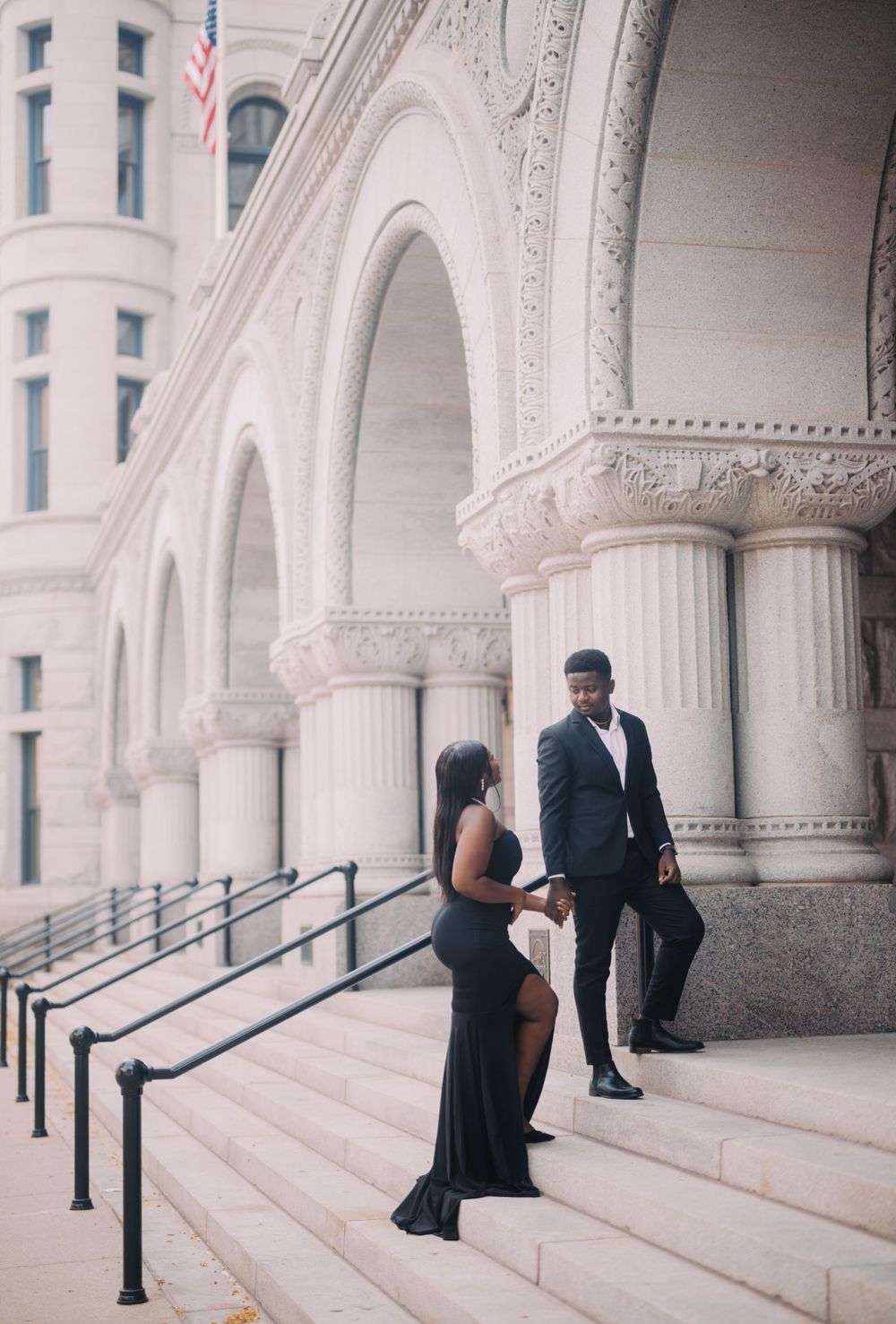 Elegant couple stands on white marble steps of a grand historic building with ornate classical columns and arches.