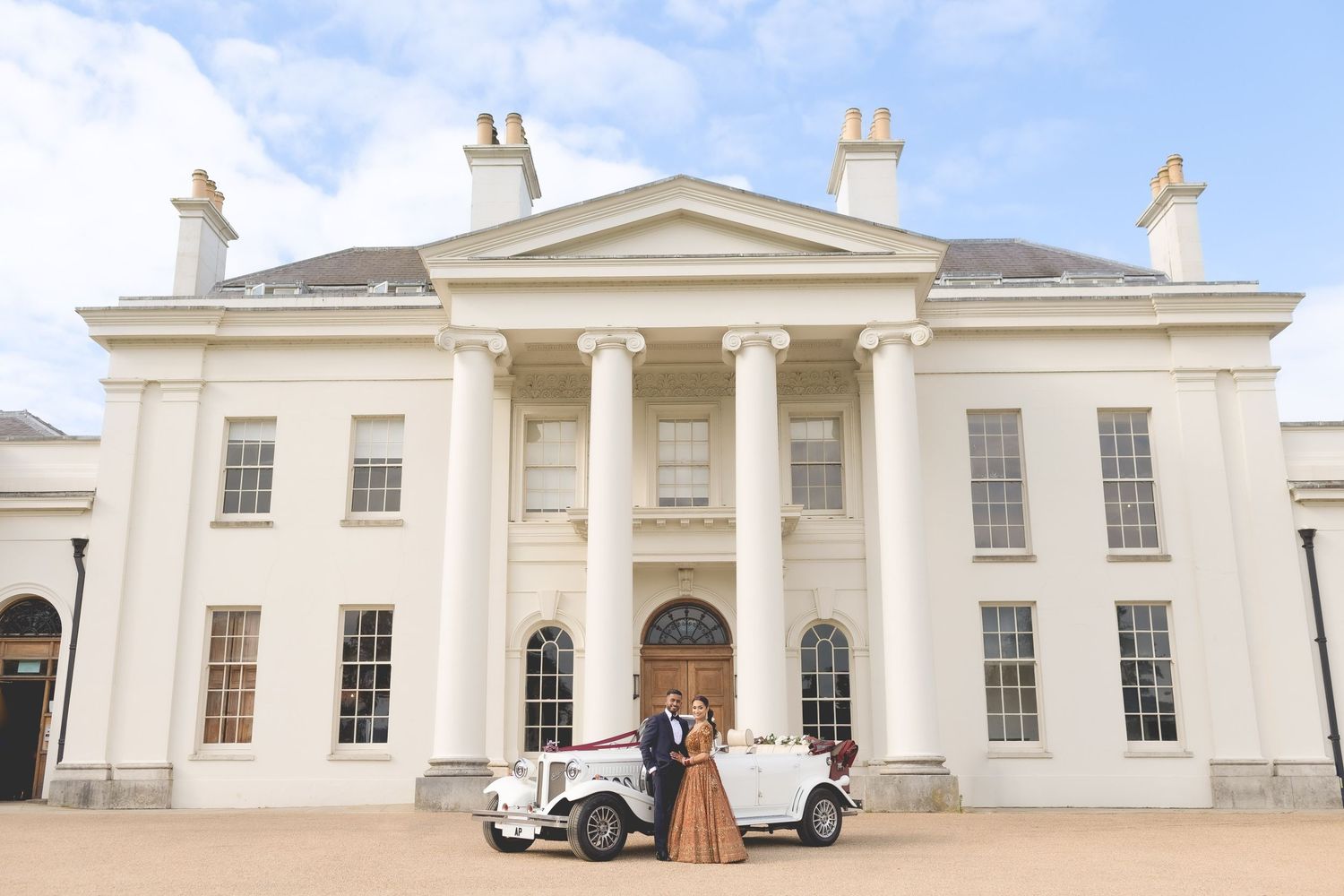 Classic vintage wedding car parked in front of grand white Georgian mansion with columns and symmetrical architecture.