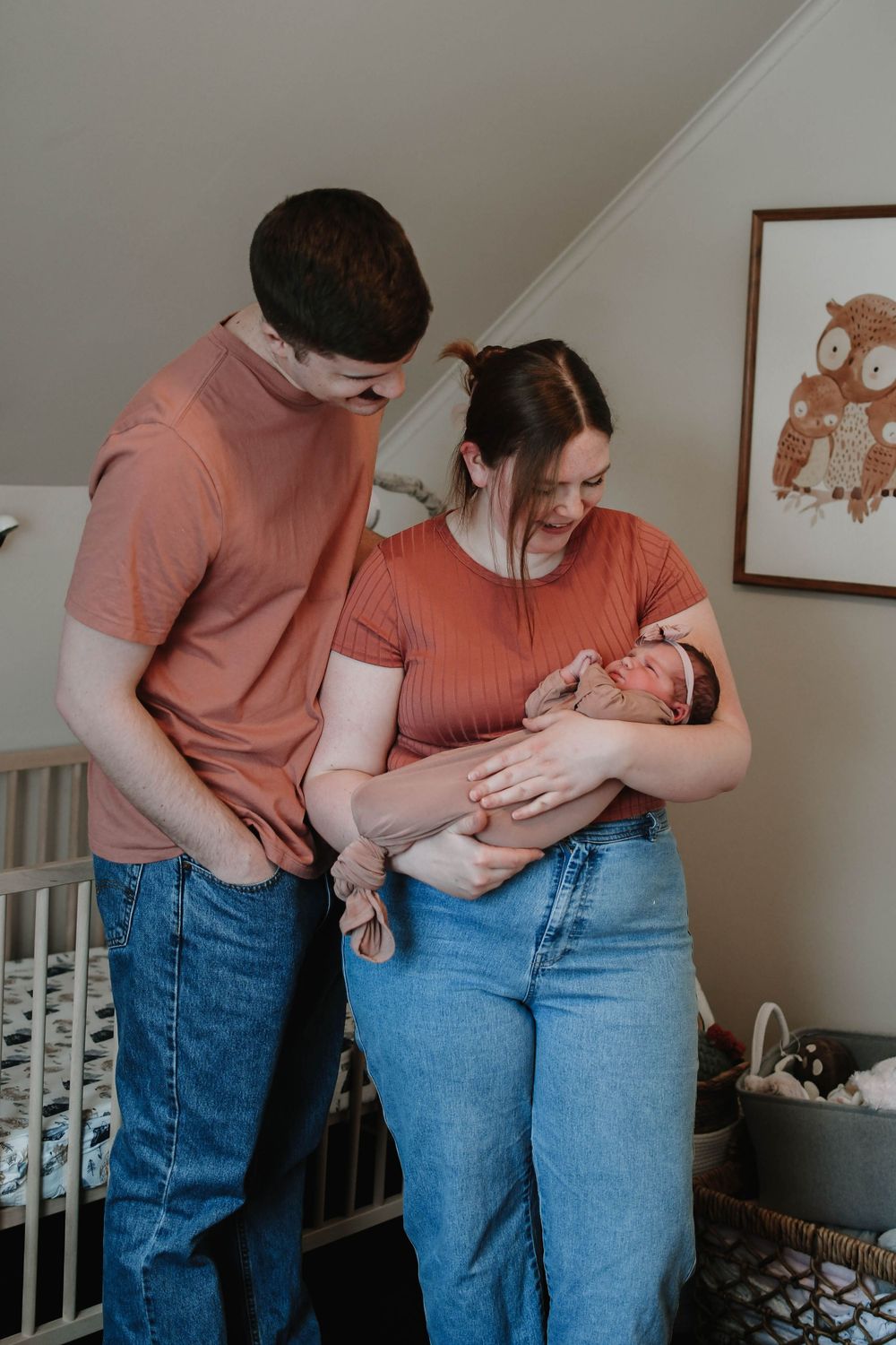 A couple in coral-colored shirts holding their newborn baby in a nursery with a gray crib and wall art.