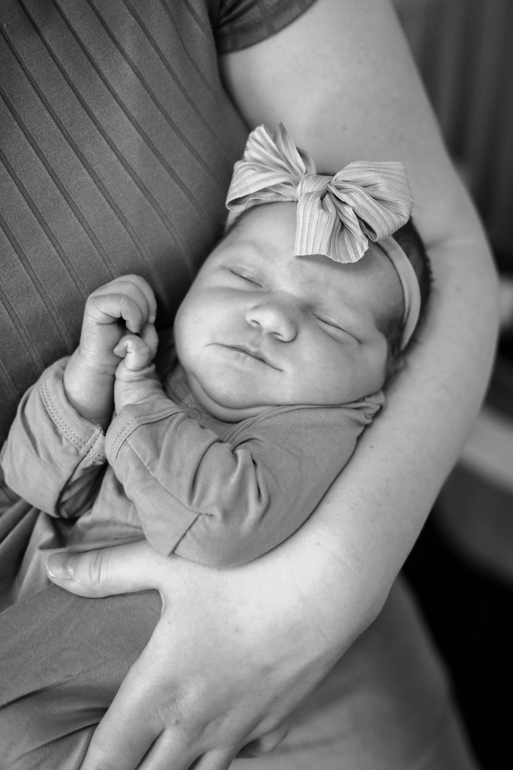 A peaceful sleeping newborn wearing a light colored bow headband rests gently in someone's arms.