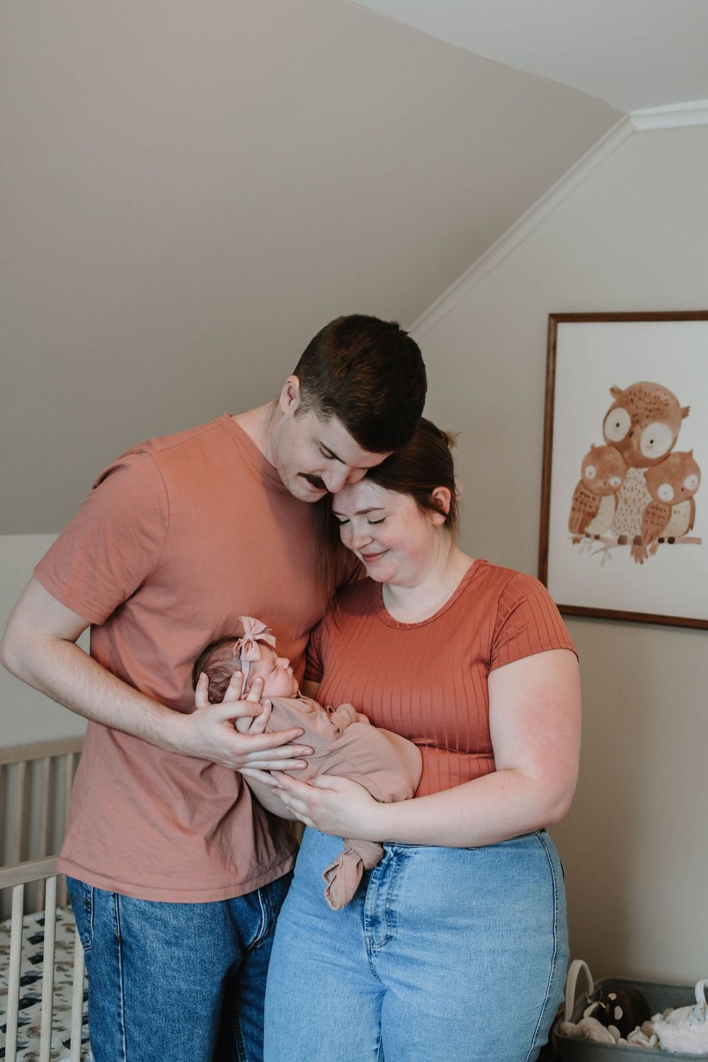 A couple in matching coral shirts stand together in a nursery with an owl print on the wall.