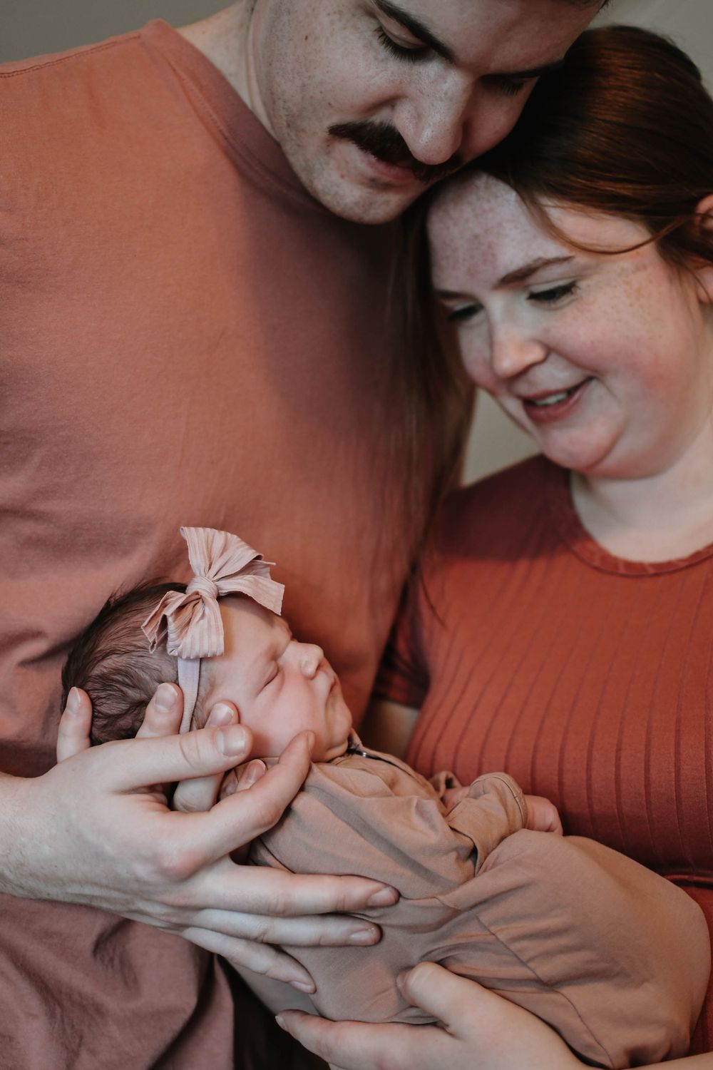 A couple in rust-colored clothing tenderly holding their sleeping newborn baby wearing a pink bow.