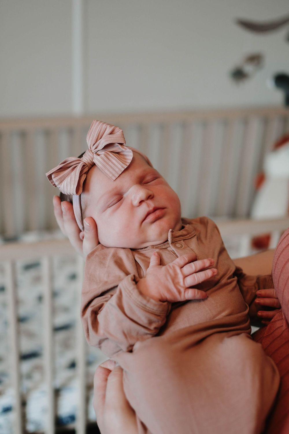 A sleeping newborn baby wearing a pink bow headband and matching pink outfit lies peacefully in a crib.
