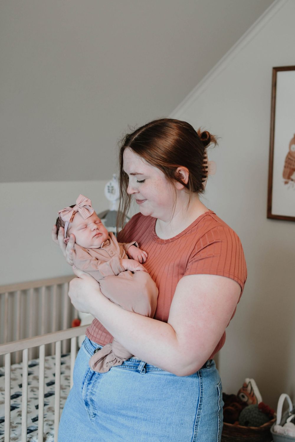 A gentle moment in a nursery as a woman in an orange shirt and jeans cradles a small baby wearing a bow.