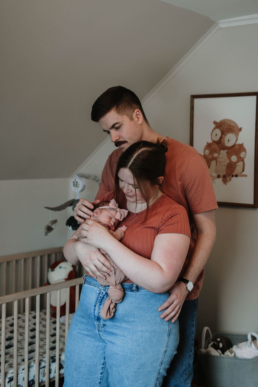 Parents holding and gazing at their newborn baby in a cozy nursery with an owl painting on the wall.