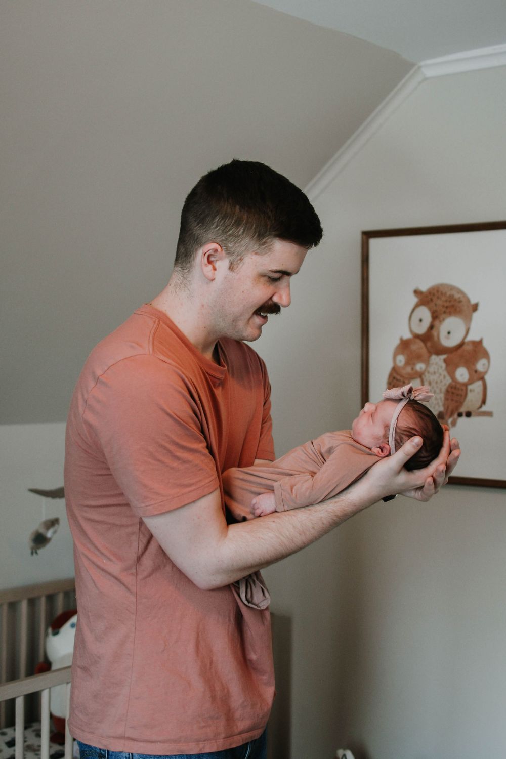 A person in a coral-colored t-shirt holding a small infant in a nursery with a teddy bear artwork on the wall.